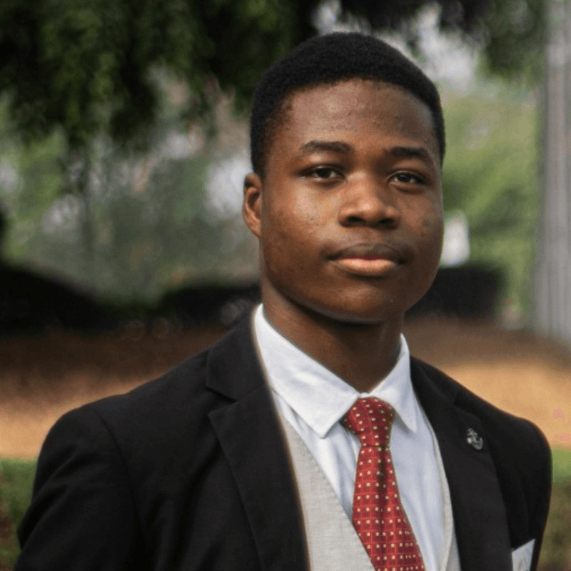 Young Black man in a formal black suit, white shirt, and red tie outdoors.